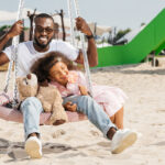 smiling african american father and daughter on spider web nest swing at amusement park
