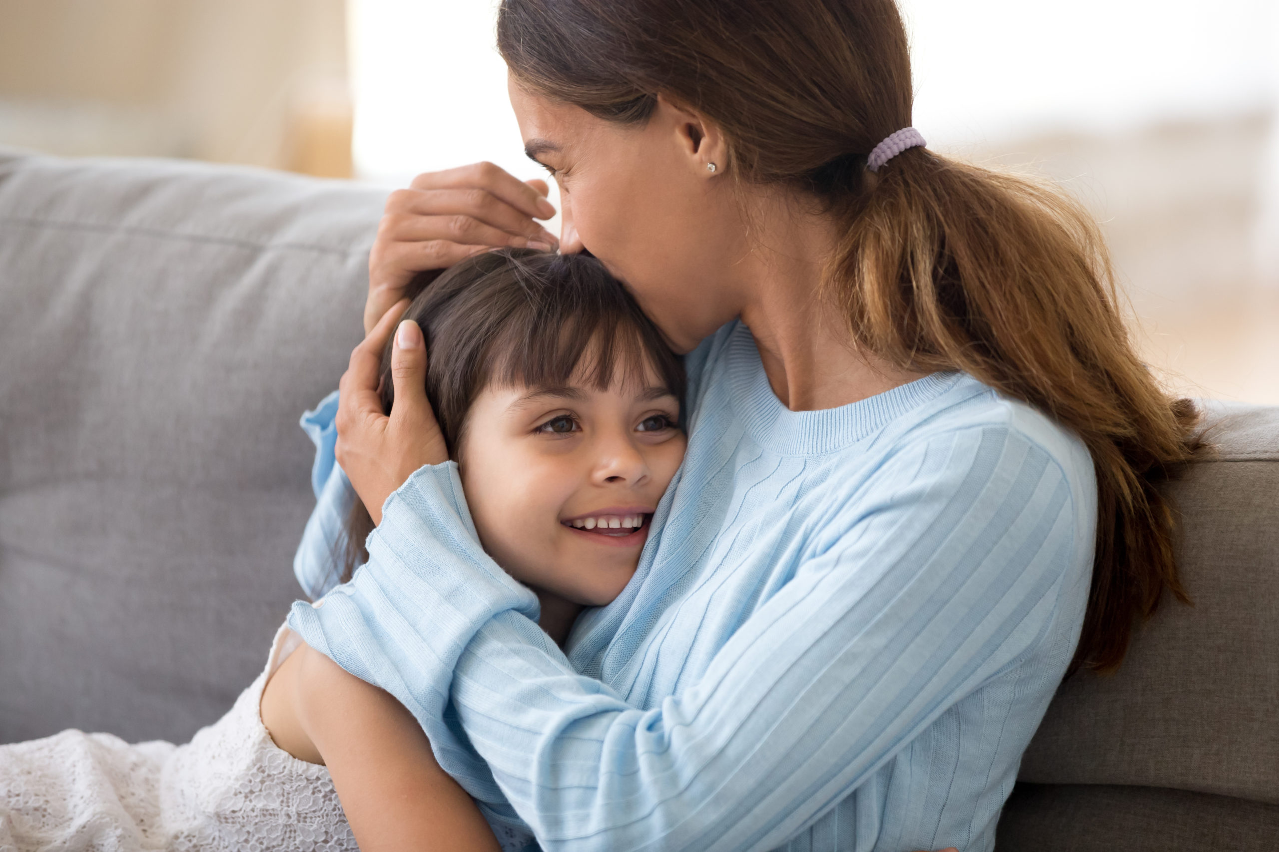 Loving mother hugging little daughter kissing kid sitting on sof