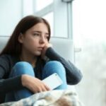 Upset teenage girl with smartphone sitting at window indoors. Space for text