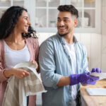 Cheerful Middle Eastern Couple Sharing Domestic Chores, Washing Dishes Together In Kitchen, Happy Millennial Arab Spouses Enjoying Making Cleaning At Home, Looking And Smiling To Each Other